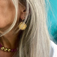 Charger l'image dans la galerie, Gros plan sur une boucle d'oreille en forme médaille soleil portée par une femme aux cheveux blanc argent en chemise blanche et portant un collier ras du cou en perles rondes or et roses.