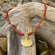 Charger l'image dans la galerie, cette photo montre un collier en perles heishi jaspe rouge et hématite or avec un pendentif africain en laiton doré en forme de 3/4 de lune posé sur un branche d'arbre vieillie par le temps