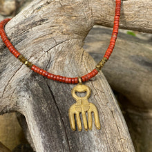 Charger l'image dans la galerie, cette photo montre un collier en perles heishi jaspe rouge et hématite or avec un pendentif africain en laiton doré en forme de main ou de fourche posé sur un branche d'arbre vieillie par le temps
