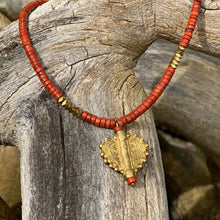 Charger l'image dans la galerie, cette photo montre un collier en perles heishi jaspe rouge et hématite or avec un pendentif africain en laiton doré en forme de coeur soleil posé sur un branche d'arbre vieillie par le temps