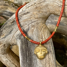 Charger l'image dans la galerie, cette photo montre un collier en perles heishi jaspe rouge et hématite or avec un pendentif africain en laiton doré en forme de tête africaine ronde posé sur un branche d'arbre vieillie par le temps