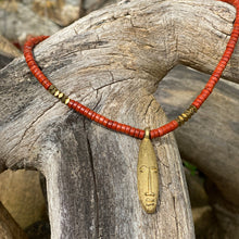 Charger l'image dans la galerie, cette photo montre un collier en perles heishi jaspe rouge et hématite or avec un pendentif africain en laiton doré en forme de tête longue posé sur un branche d'arbre vieillie par le temps