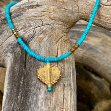 Charger l'image dans la galerie, cette photo montre un collier en perles heishi en howlite turquoise et hématite or avec un pendentif africain en laiton doré en forme de coeur soleil posé sur un branche d'arbre vieillie par le temps