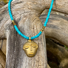Charger l'image dans la galerie, cette photo montre un collier en perles heishi en howlite turquoise et hématite or avec un pendentif africain en laiton doré en forme de tête africaine ronde posé sur un branche d'arbre vieillie par le temps