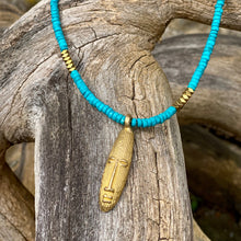 Charger l'image dans la galerie, cette photo montre un collier en perles heishi en howlite turquoise et hématite or avec un pendentif africain en laiton doré en forme de tête longue posé sur un branche d'arbre vieillie par le temps