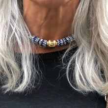 Charger l'image dans la galerie, un collier ras du cou en perles céramiques bleues du Ghana et grosse perle large dorée sur le cou d'une femme aux cheveux longs blanc argenté et pull noir