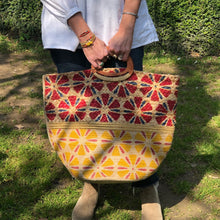 Charger l'image dans la galerie, Grand panier en jute peint à motifs rouge te jaune avec anse en bois arrondis portée main d'une femme en chemise blanche et jean dans un square parisien