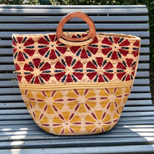 Charger l'image dans la galerie, Grand panier en jute peint à motifs rouge et jaune avec bande en lurex or et poignet en bois massif posé sur un banc