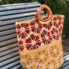 Charger l'image dans la galerie, Grand panier en jute peint à motifs rouge et jaune avec bande en lurex or et poignet en bois massif posé sur un banc