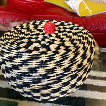 Charger l'image dans la galerie, Gros plan sur panier et son couvercle au pompon rouge en fibre végétale tressée noire et crème. Ambiance bohème chic.
