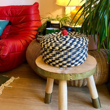 Charger l'image dans la galerie, Panier et son couvercle au pompon rouge en fibre végétale tressée noire et crème. Ambiance bohème, tabouret bois, plantes vertes et canapé design.