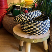 Charger l'image dans la galerie, Panier et son couvercle au pompon rouge ouvert. Fibre végétale tressée noire et crème. Ambiance bohème, tabouret bois, plantes vertes et canapé design.