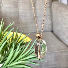 Charger l'image dans la galerie, Gros plan en transparence sur sautoir bohème grosse perle de cristal de roche et perle galet plaqué or. Plante grasse et fauteuil gris en arrière plan.