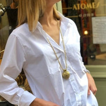Charger l'image dans la galerie, Femme blonde vêtue d'une chemise blanche simple et d'un sautoir chaine torsadée dorée avec gros pendentif africain laiton doré.Ambiance décontractée chic