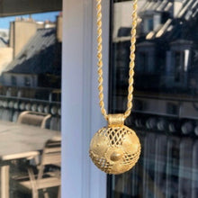 Charger l'image dans la galerie, Gros plan sur sautoir chaine et pendentif rond ajouré africain en laiton doré. Vue sur toits de Paris en arrière fond