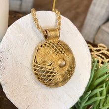 Charger l'image dans la galerie, Gros plan sur pendentif rond ajouré avec 3 petites spirales en laiton doré sur une chaine torsadée or. Posé sur un rondin de bois peint en blanc.