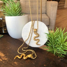 Charger l'image dans la galerie, 2 sautoirs chaine torsadée dorée avec pendentif serpent laiton doré posés sur une table en vois foncée et pots avec plantes grasses. Ambiance ethnique chic