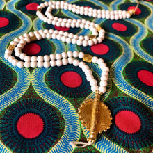 Charger l'image dans la galerie, Long sautoir en perles de bois blanc et laiton dorées avec pendentif soleil en laiton doré. Présentation sur tissu africain couleurs vives