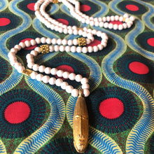 Charger l'image dans la galerie, Long sautoir en perles de bois blanc et laiton dorées avec pendentif tête longue africaine en laiton doré. Présentation sur tissu africain couleurs vives