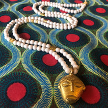 Charger l'image dans la galerie, Long sautoir en perles de bois blanc et laiton dorées avec pendentif tête ronde africaine en laiton doré. Présentation sur tissu africain couleurs vives