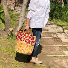 Charger l'image dans la galerie, Grand panier en jute peint à motifs rouge te jaune avec anse en bois arrondis portée main d'une femme en chemise blanche et jean dans un square parisien