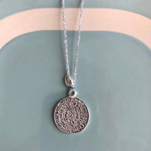 Charger l'image dans la galerie, gros plan sur un sautoir talisman bohème médaille gravée argent montée sur fil argent et associée à une perle galet plaqué argent. Fond bleu vintage et blanc.