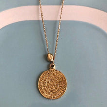 Charger l'image dans la galerie, gros plan sur un sautoir talisman bohème médaille gravée or montée sur fil or et associée à une perle galet dorée à l'or fin. Fond bleu vintage et blanc.