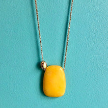Charger l'image dans la galerie, Gros plan sur sautoir talisman bohème avec grosse perle plate en agate jaune et galet doré à l'or fin sur fil doré. Fond bleu turquoise.