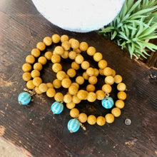 Charger l&#39;image dans la galerie, Présentation d&#39;un ensemble de plusieurs bracelets de grosses perles en Jaspe Mokaite poli jaune indien associées à une perle pampille en céramique bleue clair sur une planche de bois vieilli. . Ambiance exotique.
