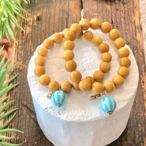 Présentation de 2  bracelets de grosses perles en Jaspe Mokaite poli jaune indien associées à une perle pampille en céramique bleue clair sur rondin de  bois blanc et planche de bois vieilli. . Ambiance exotique.