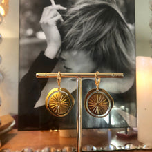 Charger l&#39;image dans la galerie, Paire de boucles d&#39;oreille médaille soleil dorée forme ajourée et design sur un portant doré avec en fond portrait de femme fumant dans un cadre miroir

