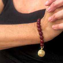Charger l&#39;image dans la galerie, Bracelet en perles bordeaux rondes avec une brloque africaine en laiton doré sur poignet femme et fond noir
