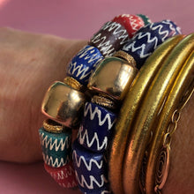 Charger l&#39;image dans la galerie, Gros plan sur 2 bracelets en perles bleues ou multicolores du Ghana et perle large et dorée mélangées avec des bracelets joncs bouddhiste or sur poignet
