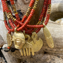 Charger l&#39;image dans la galerie, Gros plan sur colliers Africa Juju en perles heishi jaspe rouge et hématite or avec différents pendentifs africains en laiton doré. En fond écorce arbre veilli par le temps

