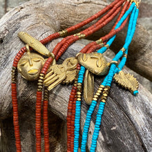 Charger l&#39;image dans la galerie, Ensemble de plusieurs colliers Africa juju en perles heishi jaspe rouge, howlite turquoise et hématite or avec pendentifs africains en laiton doré posés sur arbre à l&#39;écorce veillie par le temps
