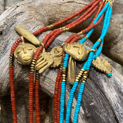 Ensemble de plusieurs colliers Africa juju en perles heishi jaspe rouge, howlite turquoise et hématite or avec pendentifs africains en laiton doré posés sur arbre à l'écorce veillie par le temps