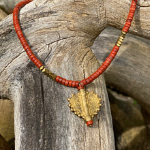 Charger l&#39;image dans la galerie, cette photo montre un collier en perles heishi jaspe rouge et hématite or avec un pendentif africain en laiton doré en forme de coeur soleil posé sur un branche d&#39;arbre vieillie par le temps
