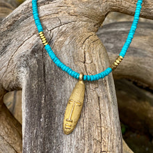 Charger l&#39;image dans la galerie, cette photo montre un collier en perles heishi en howlite turquoise et hématite or avec un pendentif africain en laiton doré en forme de tête longue posé sur un branche d&#39;arbre vieillie par le temps
