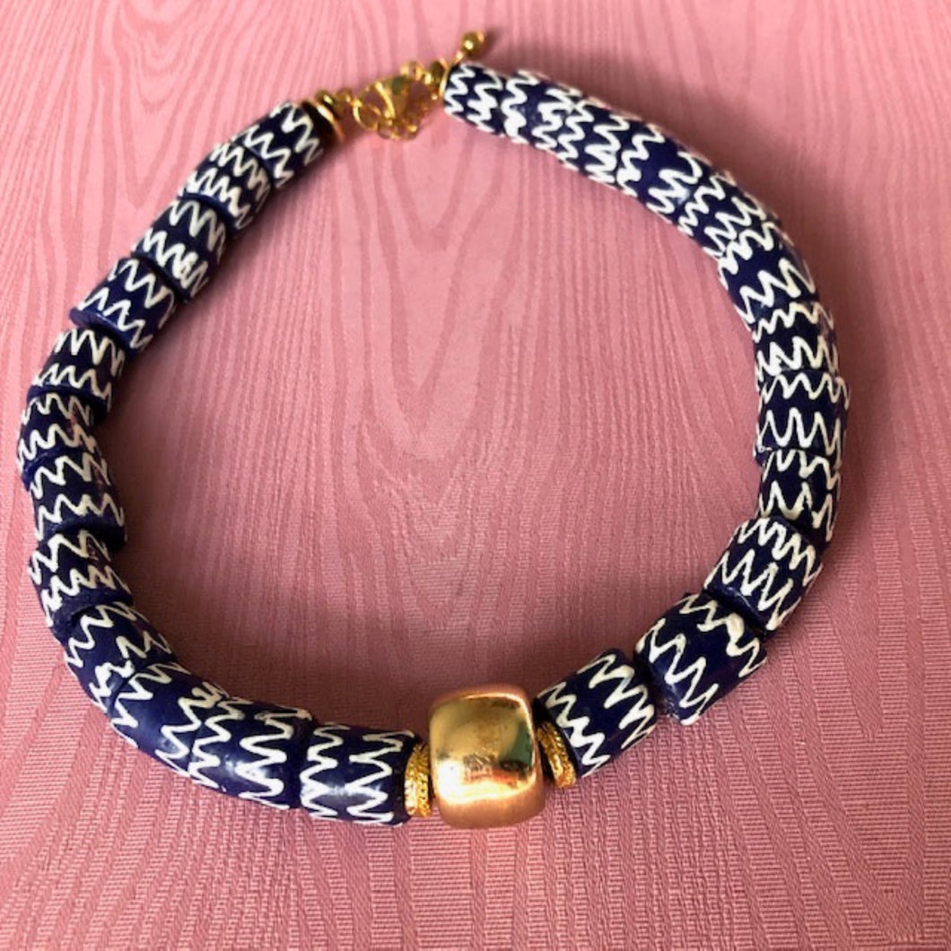 un collier ras du cou en perles céramiques bleues du Ghana et grosse perle large dorée sur un fond de tissu rose