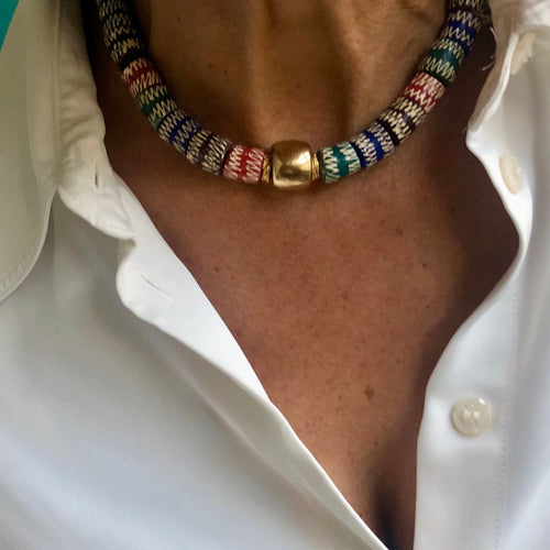 un collier ras du cou en perles céramiques multicolores du Ghana et grosse perle large dorée sur le cou d'une femme aux cheveux longs blanc argenté et chemise blanche