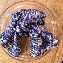 Charger l&#39;image dans la galerie, Grande écharpe en soie bleu et violet, nouée et posée sur plateau de verre et table en bois blond
