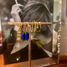 Charger l&#39;image dans la galerie, Plusieurs petites boucles d&#39;oreille dont une avec une perle facettée en Lapis Lazuli sur un portant doré avec en fond portrait de femme fumant dans un cadre miroir
