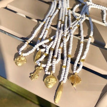 Charger l&#39;image dans la galerie, Présentation d&#39;une série de 6 sautoirs en perles de bois blanc avec pendentifs africains en laiton doré.Ambiance ensoleillée sur chaise métal jardin du Luxembourg
