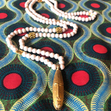 Charger l&#39;image dans la galerie, Long sautoir en perles de bois blanc et laiton dorées avec pendentif tête longue africaine en laiton doré. Présentation sur tissu africain couleurs vives

