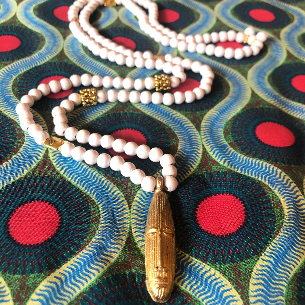 Long sautoir en perles de bois blanc et laiton dorées avec pendentif tête longue africaine en laiton doré. Présentation sur tissu africain couleurs vives
