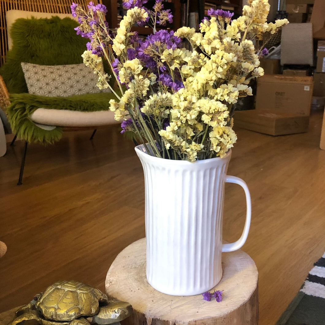 Vase pichet en porcelaine blanche avec un bouquet de fleurs séchées jaune et violet posée sur un tronc d'arbre dans un intérieur bohème