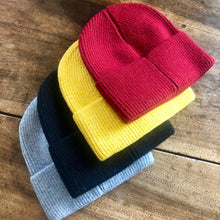 Charger l&#39;image dans la galerie, 4 bonnets en laine unis, rouge, jaune, marine et bleu ciel sur fond en bois.
