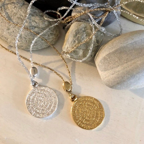 plan sérré sur 2 sautoirs talimans bohèmes sur forme médaille gravée or et argent posés à plat à coté de cailloux de différentes formes texture.