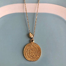 Charger l&#39;image dans la galerie, gros plan sur un sautoir talisman bohème médaille gravée or montée sur fil or et associée à une perle galet  dorée à l&#39;or fin. Fond bleu vintage et blanc.
