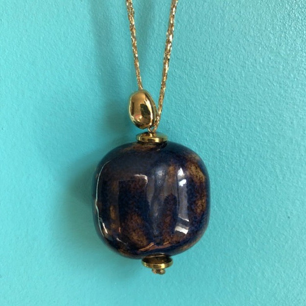 Gros plan sur sautoir talisman bohème avec grosse perle en céramique marron bleue et galet doré à l'or fin sur fil doré. Fond bleu turquoise.